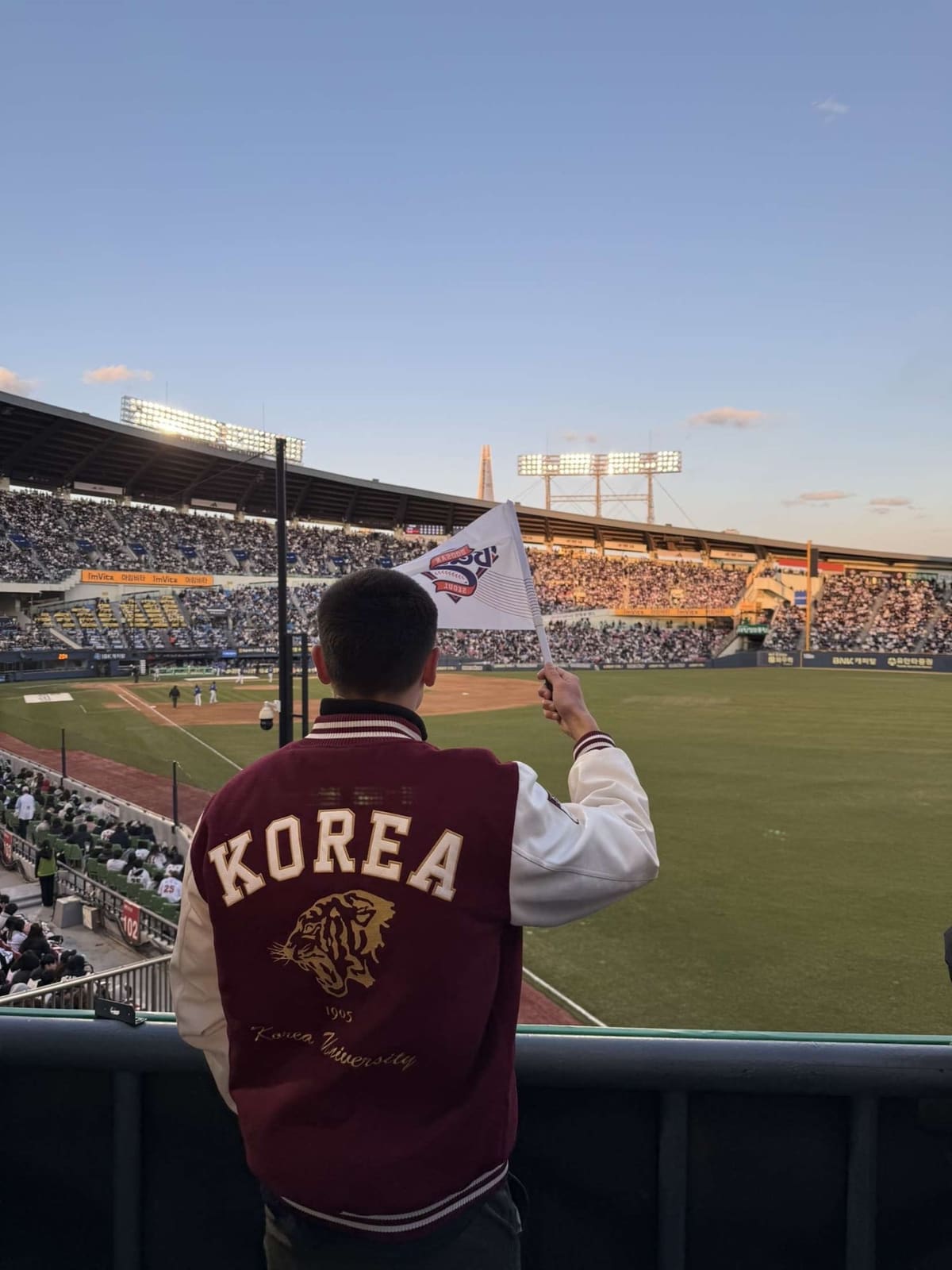 William Stoops, seen from behind in a Korea University jacket, waves a flag in the stands of a Seoul baseball stadium